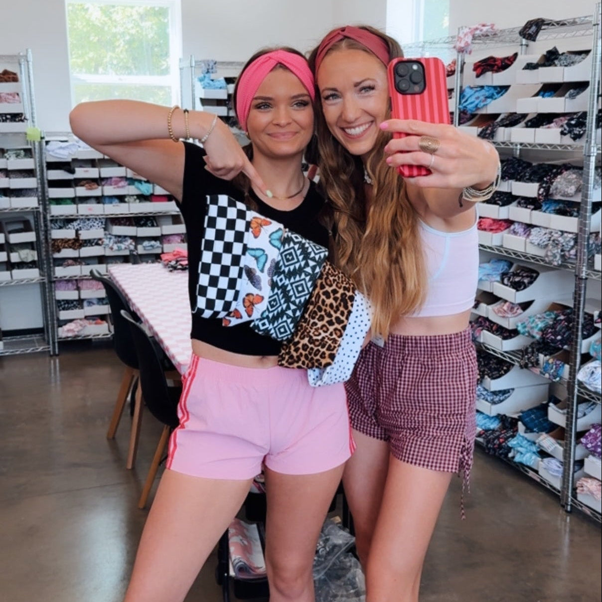 Two women taking a selfie in a headband store. The woman on the left wears a black graphic crop top, a pink headband, and pink shorts.