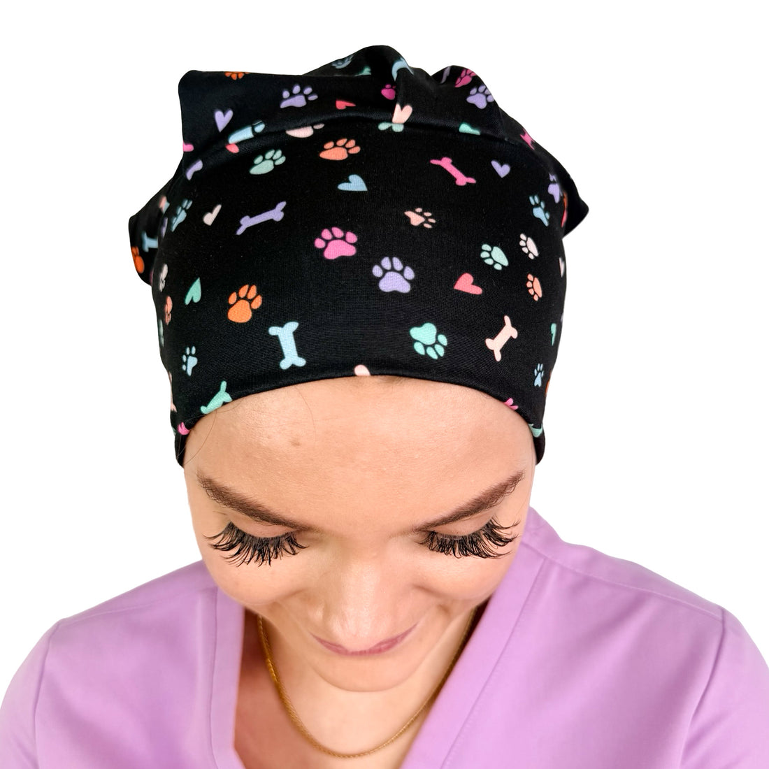 A woman wearing a black scrub cap with a colorful pattern of dog bones, paw prints, and hearts, paired with lavender scrubs against a white background.
